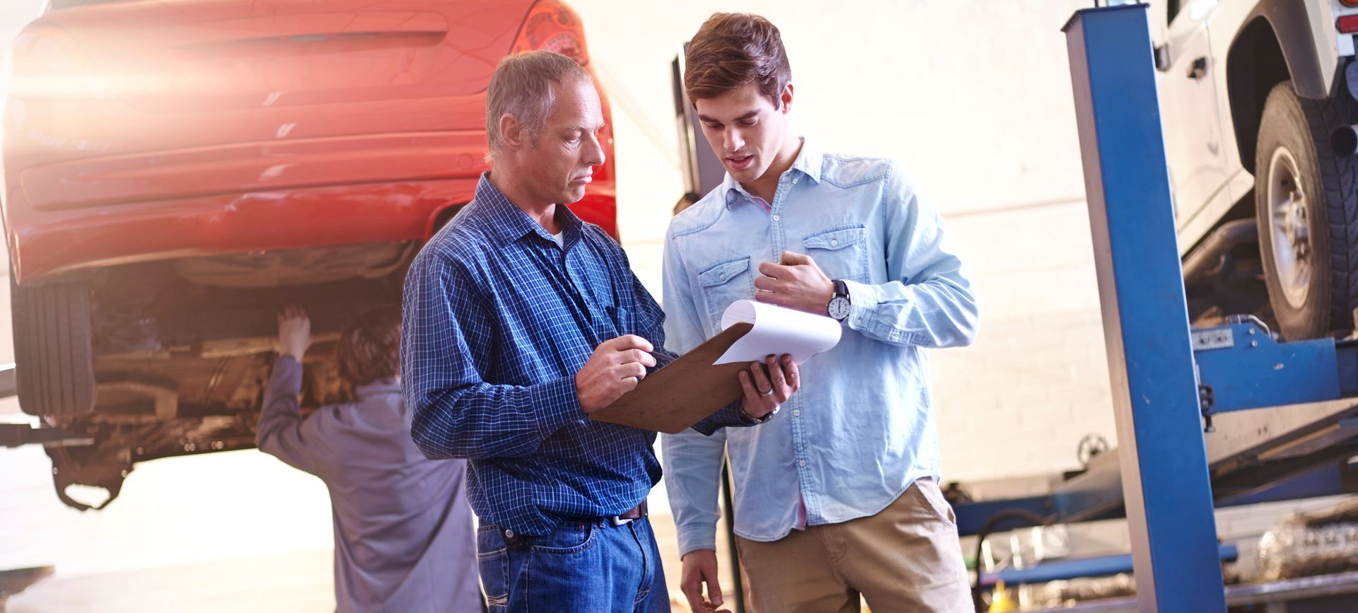 Two men reviewing paperwork near a car on a lift in a garage; a mechanic works below the car.