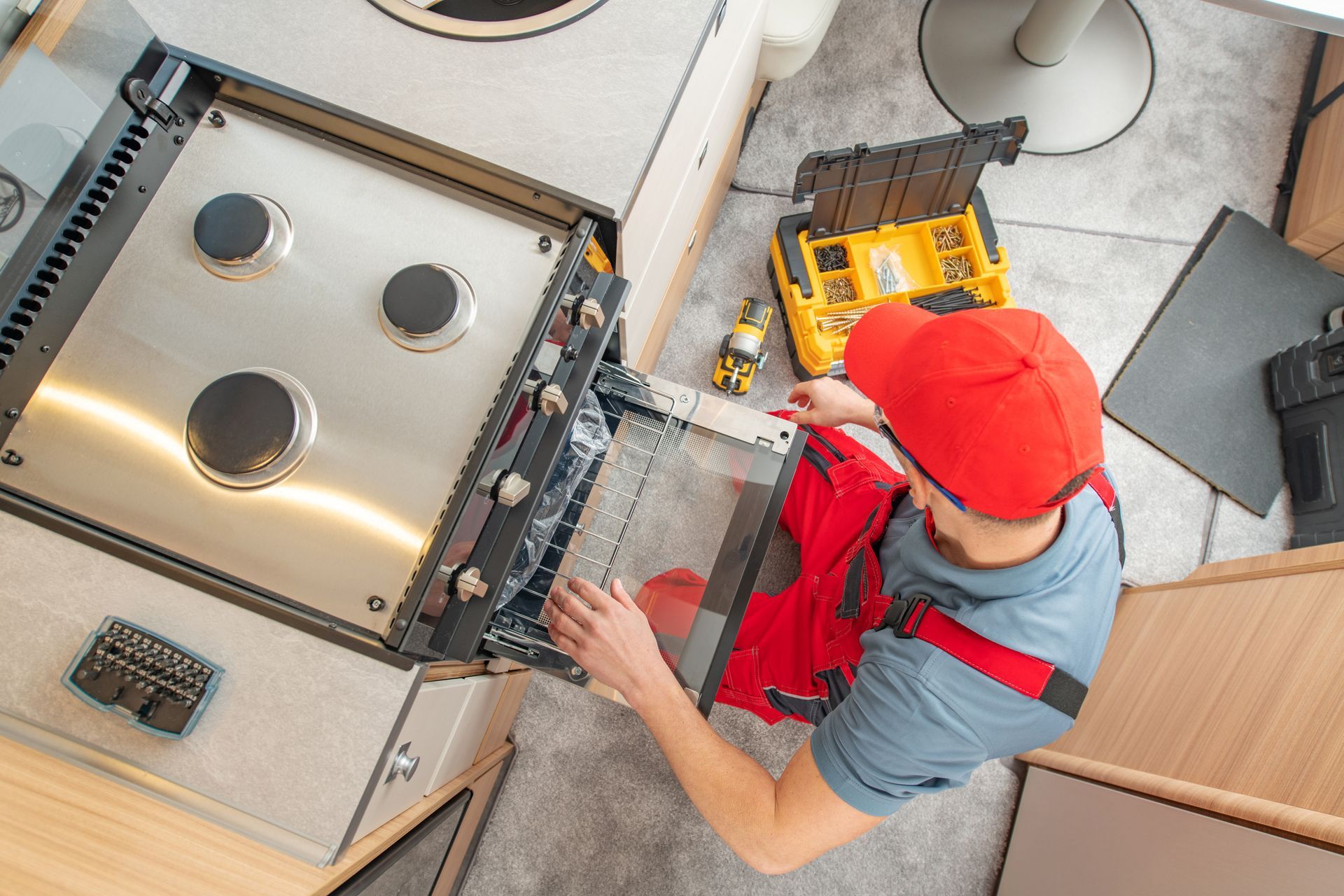 A technician in a red cap and uniform repairs a stainless-steel oven inside a camper or kitchen.