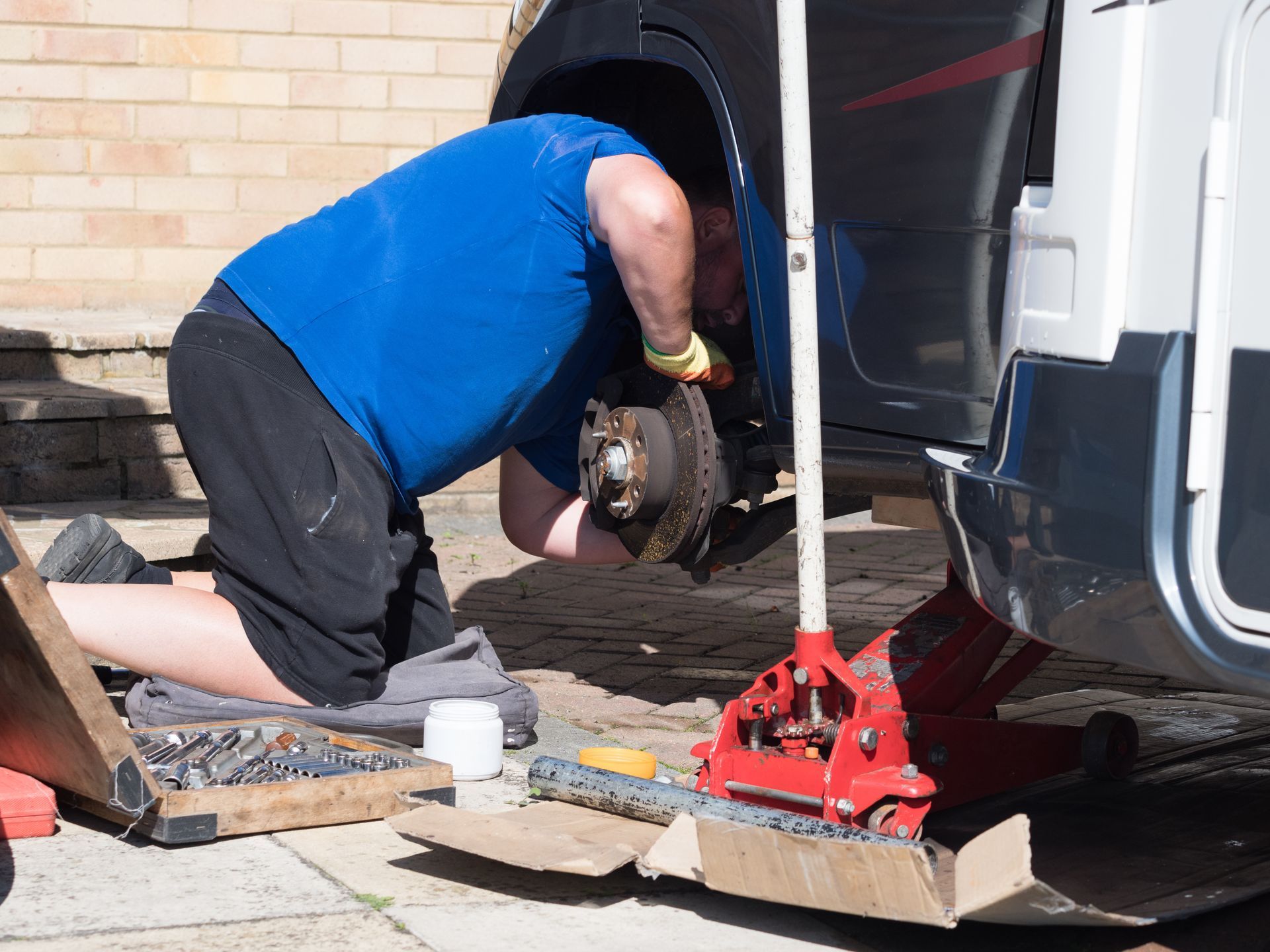 A person wearing a blue shirt and yellow gloves works on the wheel hub of a vehicle raised by a red hydraulic jack.