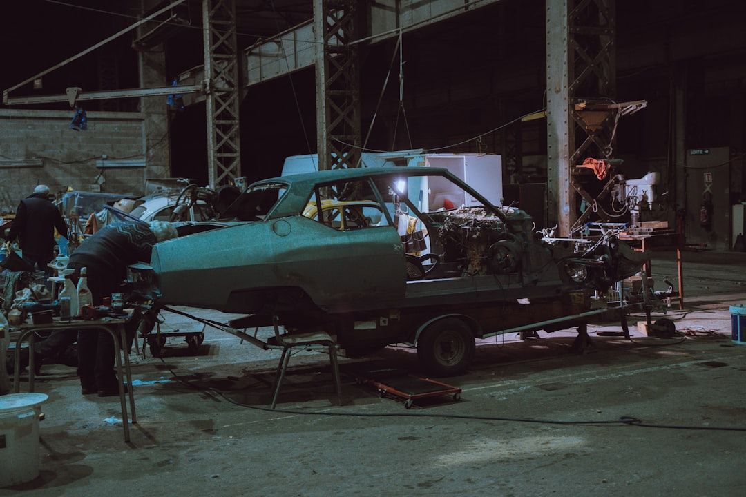 A green car body on a trailer inside a building. Workers are seen near the vehicle at night.