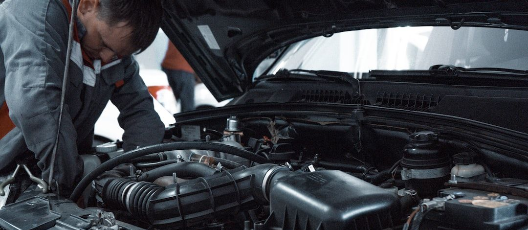 Mechanic in blue jumpsuit works on a car engine with the hood open, indoor setting.