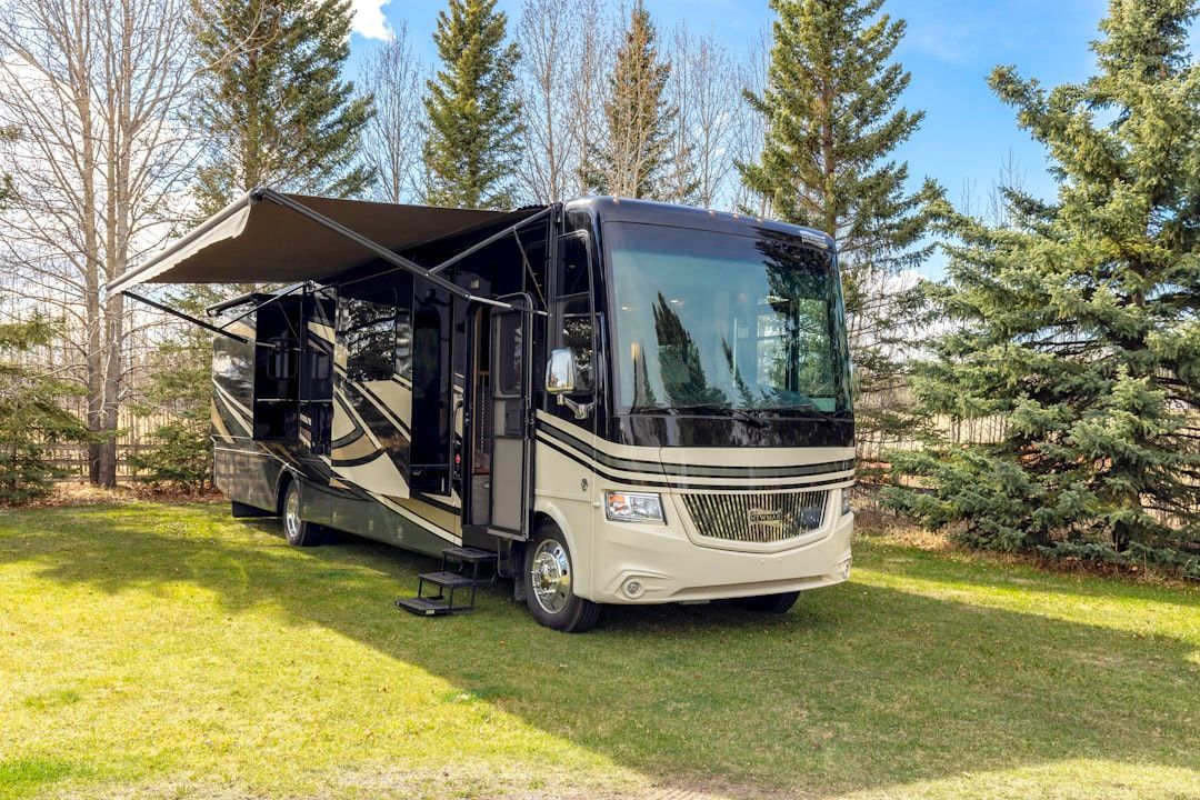 A modern tan and black motorhome with its awning extended, parked on a grassy lot surrounded by tall pine trees.