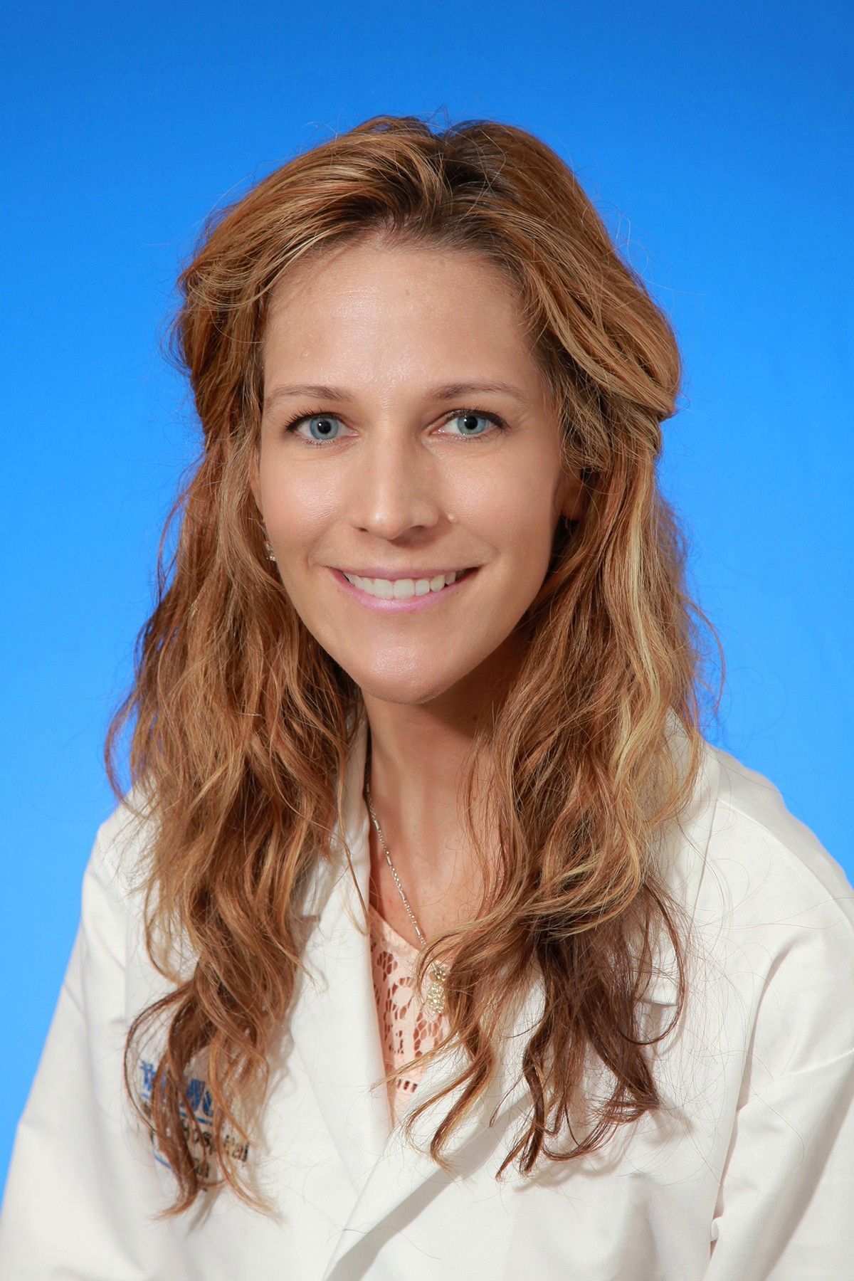 A woman in a white lab coat is smiling in front of a blue background.