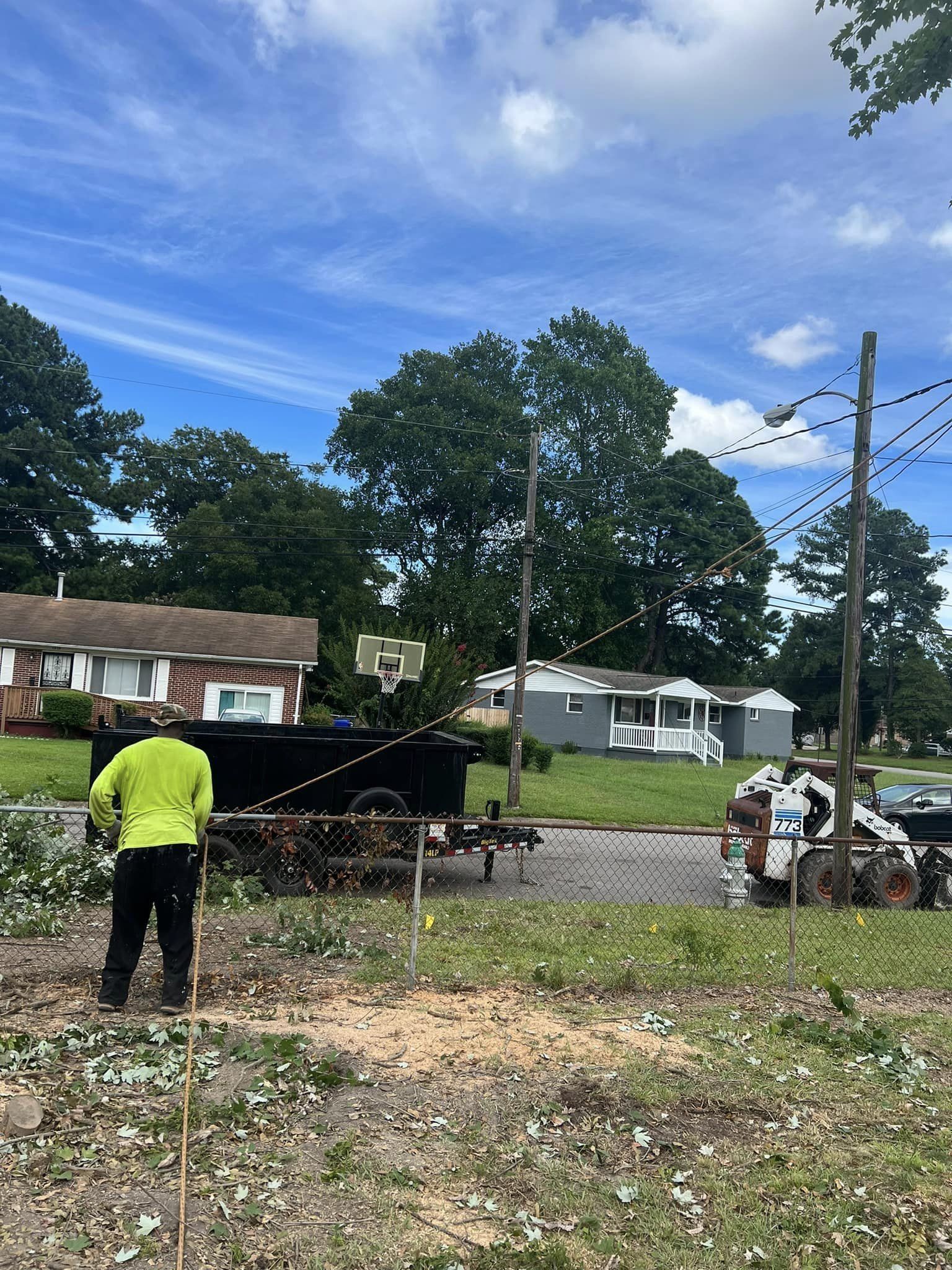 A person in a bright yellow shirt stands near a chain-link fence by a black dump trailer and a skid steer at a yard site.