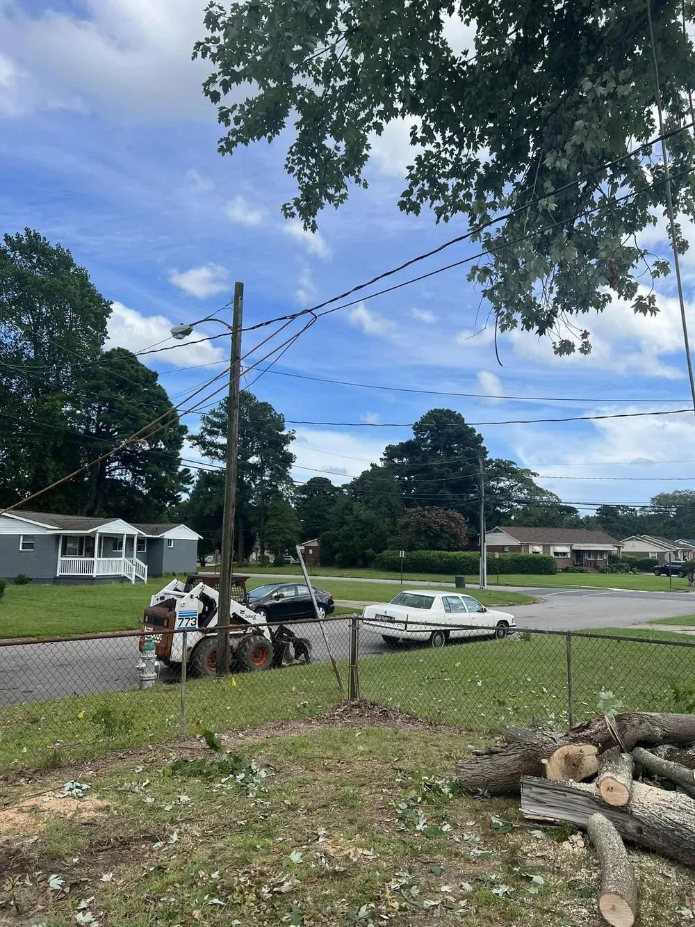 A skid steer parked on a gravel roadside near a residential house, trees, and a parked car under a partly cloudy sky.