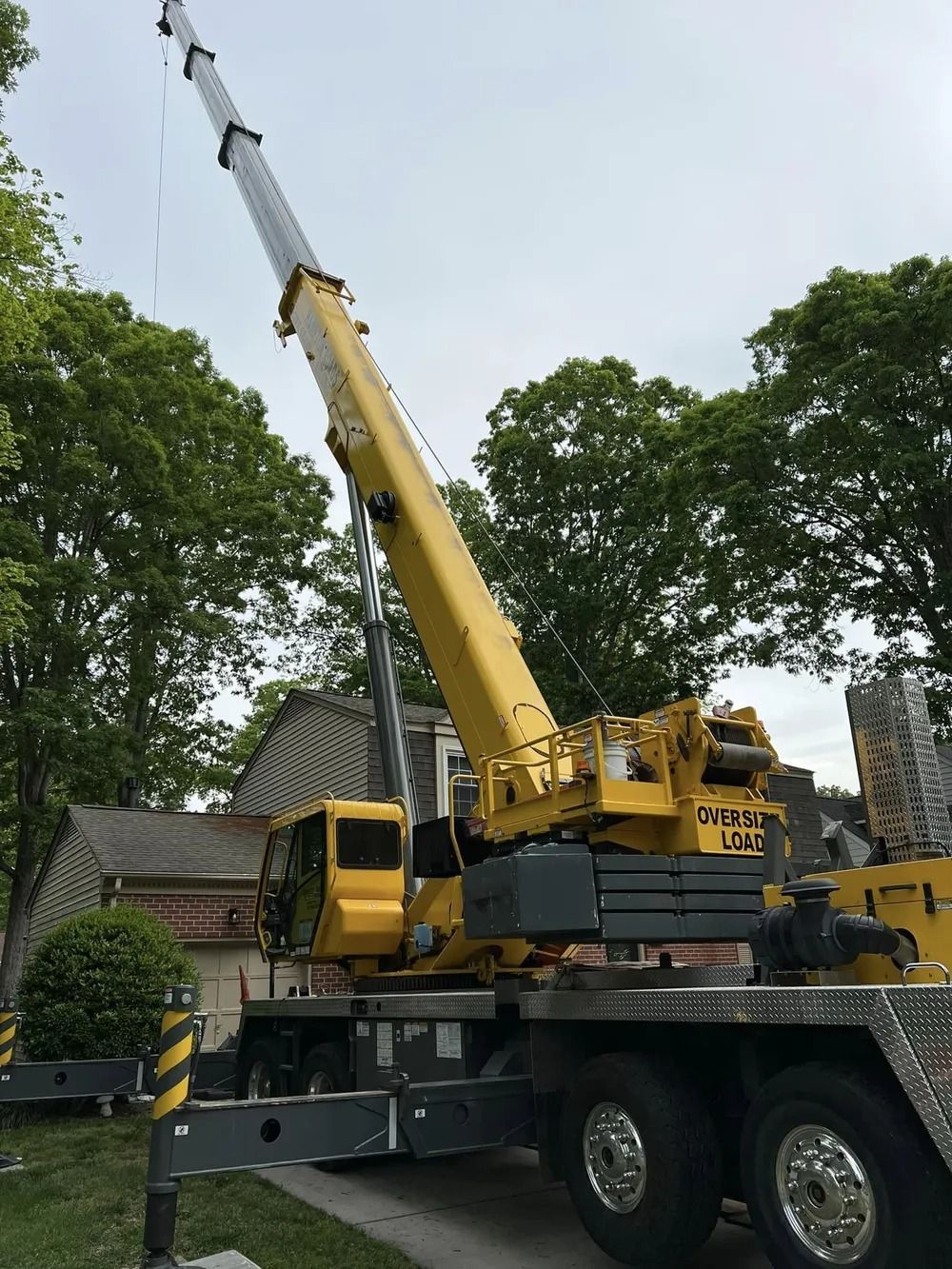 A yellow mobile crane with an extended boom is positioned on a residential street in front of a house and trees.