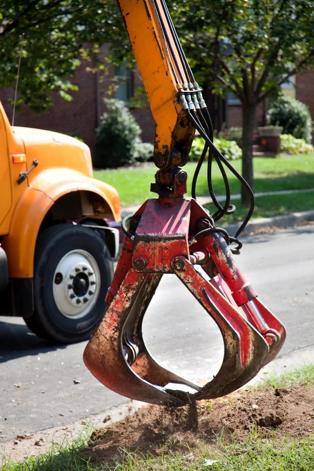 A heavy-duty orange and red hydraulic grapple attached to a truck arm, positioned above a small patch of dirt.