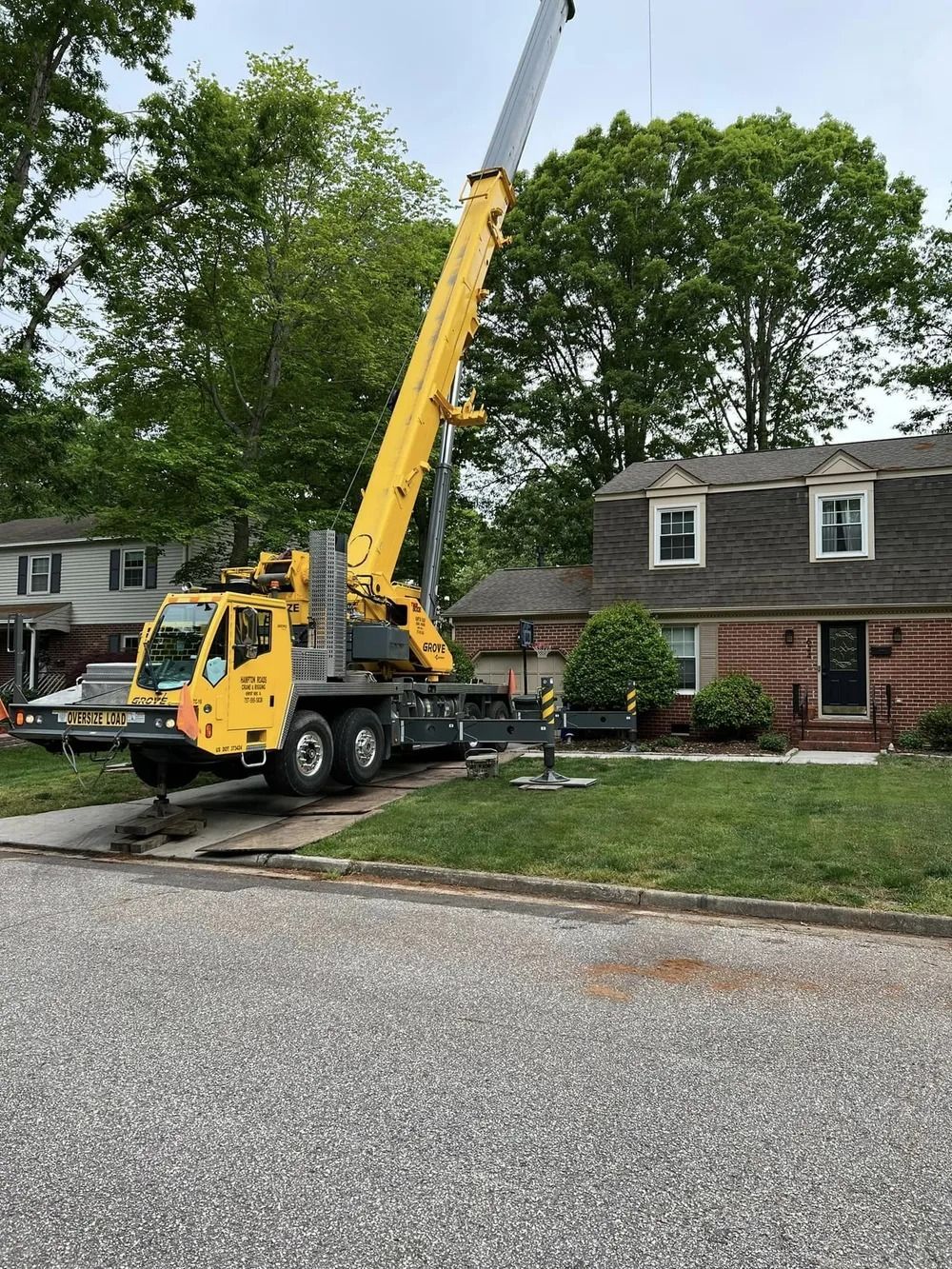 A bright yellow crane parked on a suburban lawn, its boom extended high above a two-story brick house.