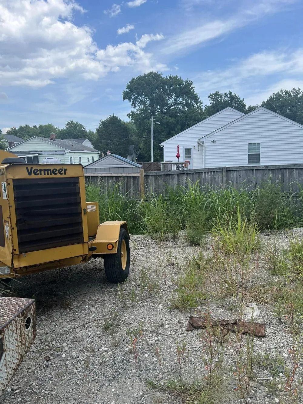 A yellow Vermeer industrial machine sits on a gravel lot in front of a wooden fence and suburban houses under a blue sky.