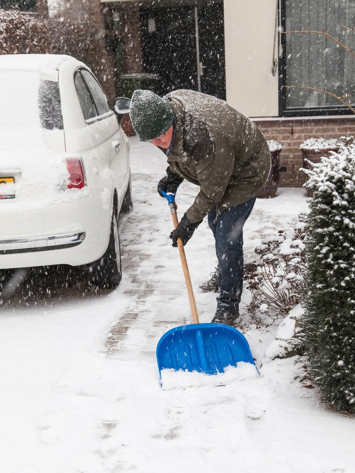 A person in a dark coat and hat shovels snow from a residential driveway next to a white car during a snowstorm.