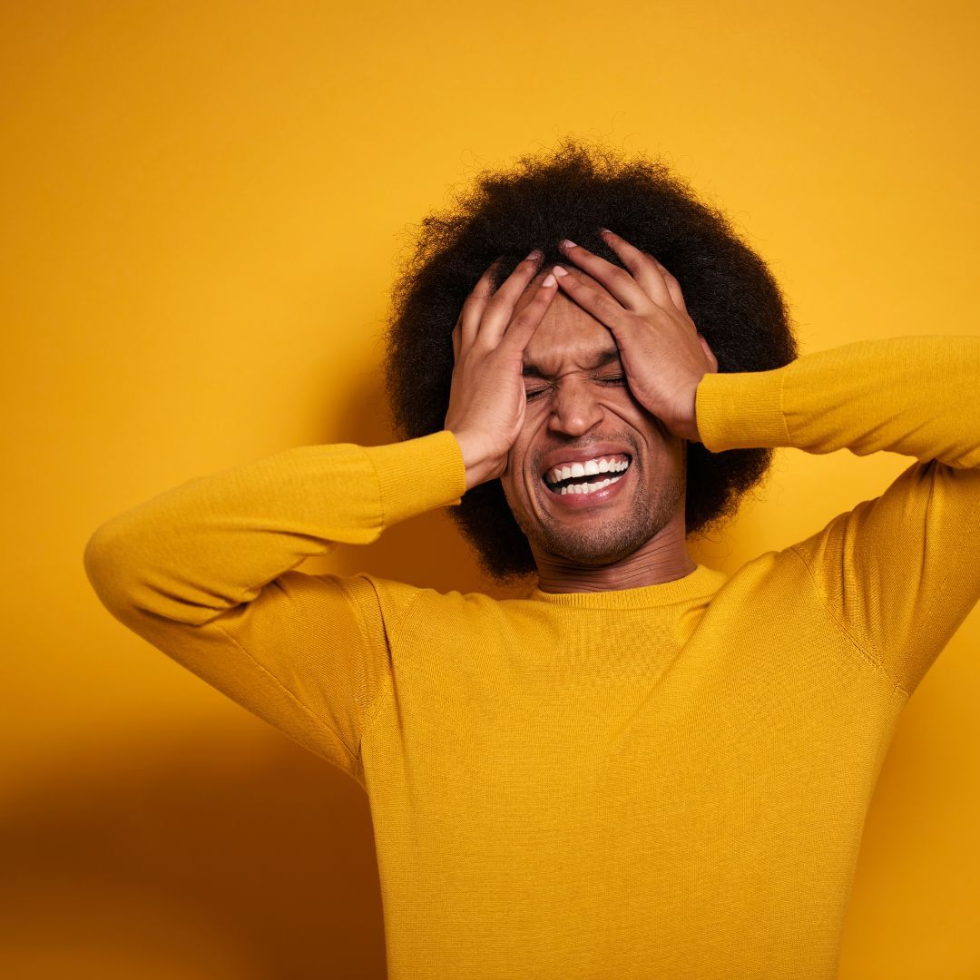 A person with curly hair wearing a yellow sweater, holding their head with both hands while laughing against a yellow wall.