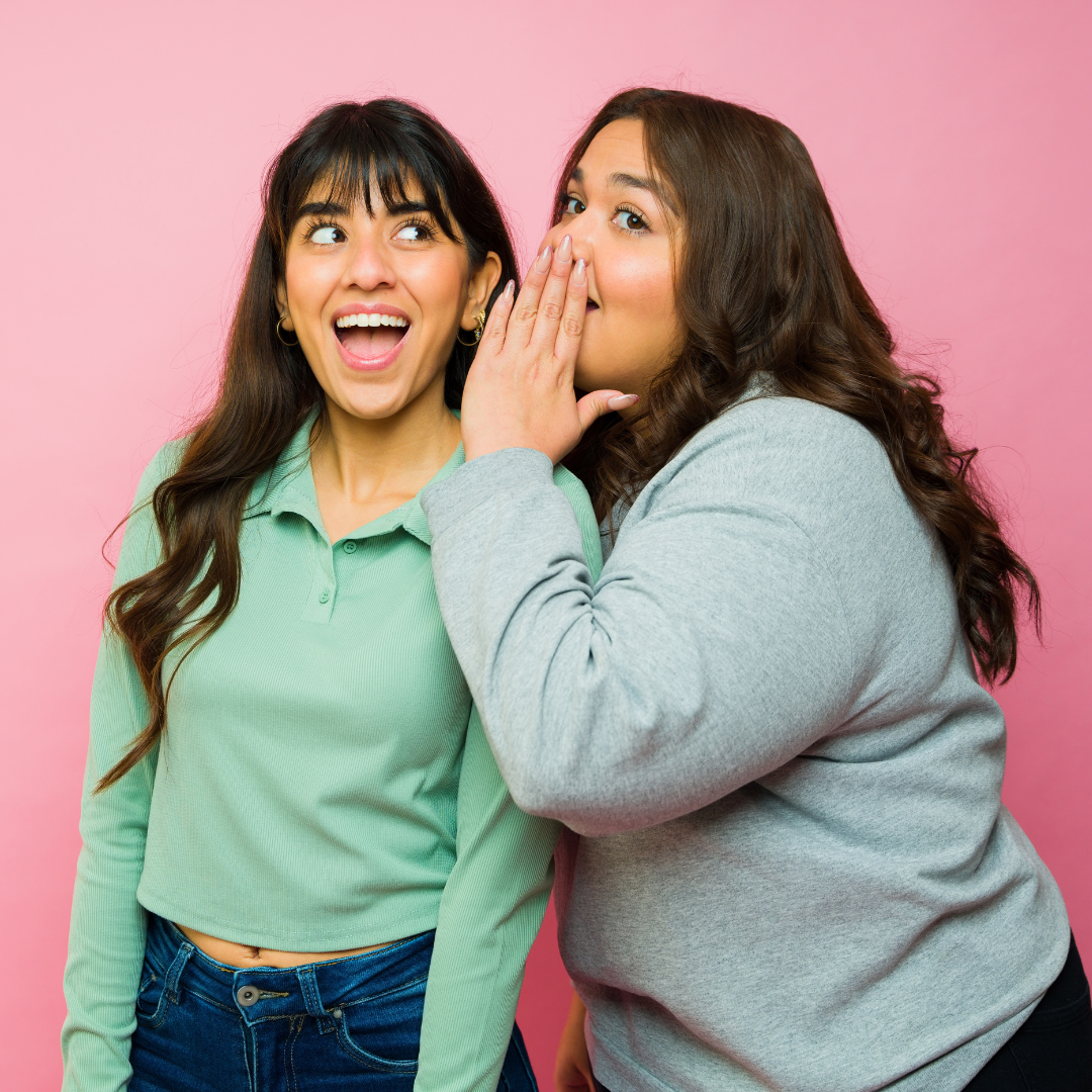 Woman whispering into another woman's ear, both smiling, pink background.