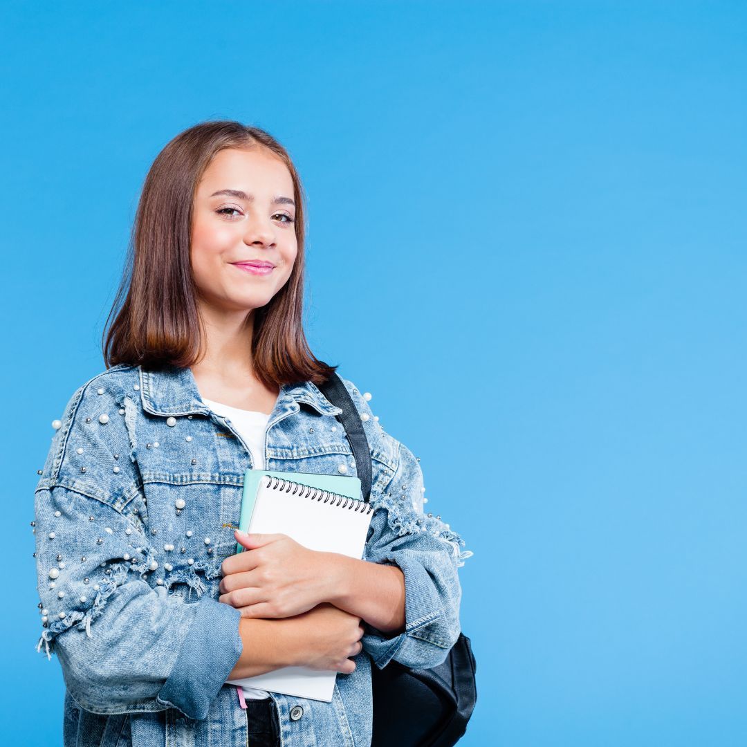 Teenager with a notebook and backpack smiles against a blue background, wearing a jean jacket.