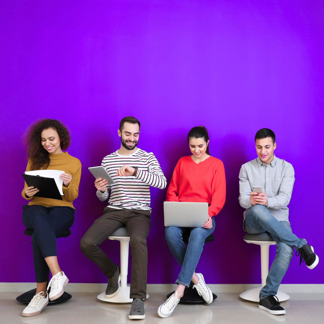 Four people sit on stools against a purple wall, using a clipboard, tablet, laptop, and phone.