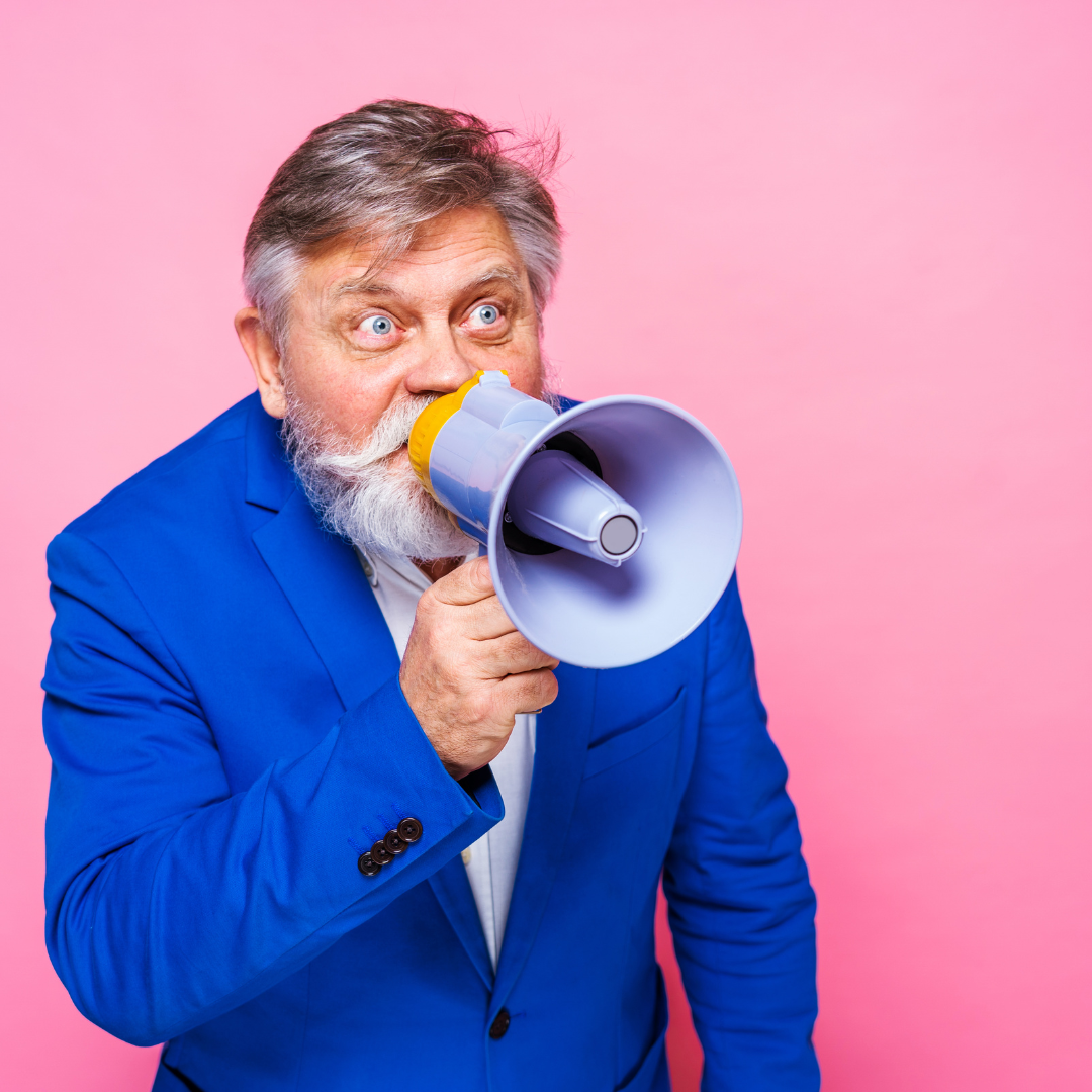 A person with a beard in a bright blue suit holds a megaphone to their mouth against a solid pink background.