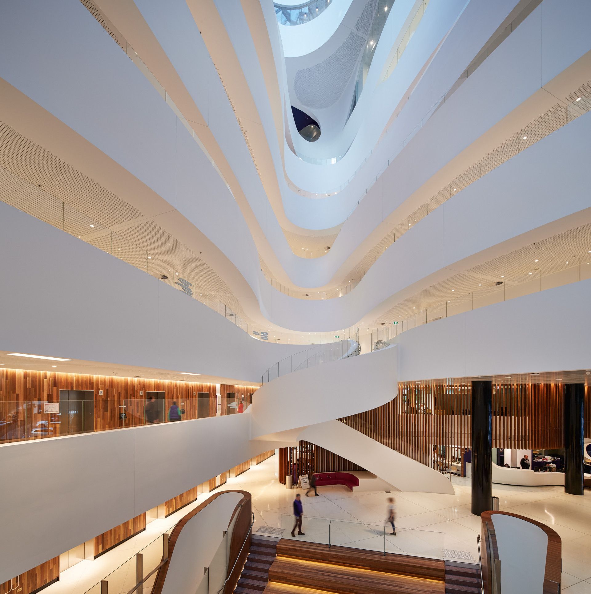 Interior view of a modern building with curved white balconies and a grand staircase; people walk on the floors.