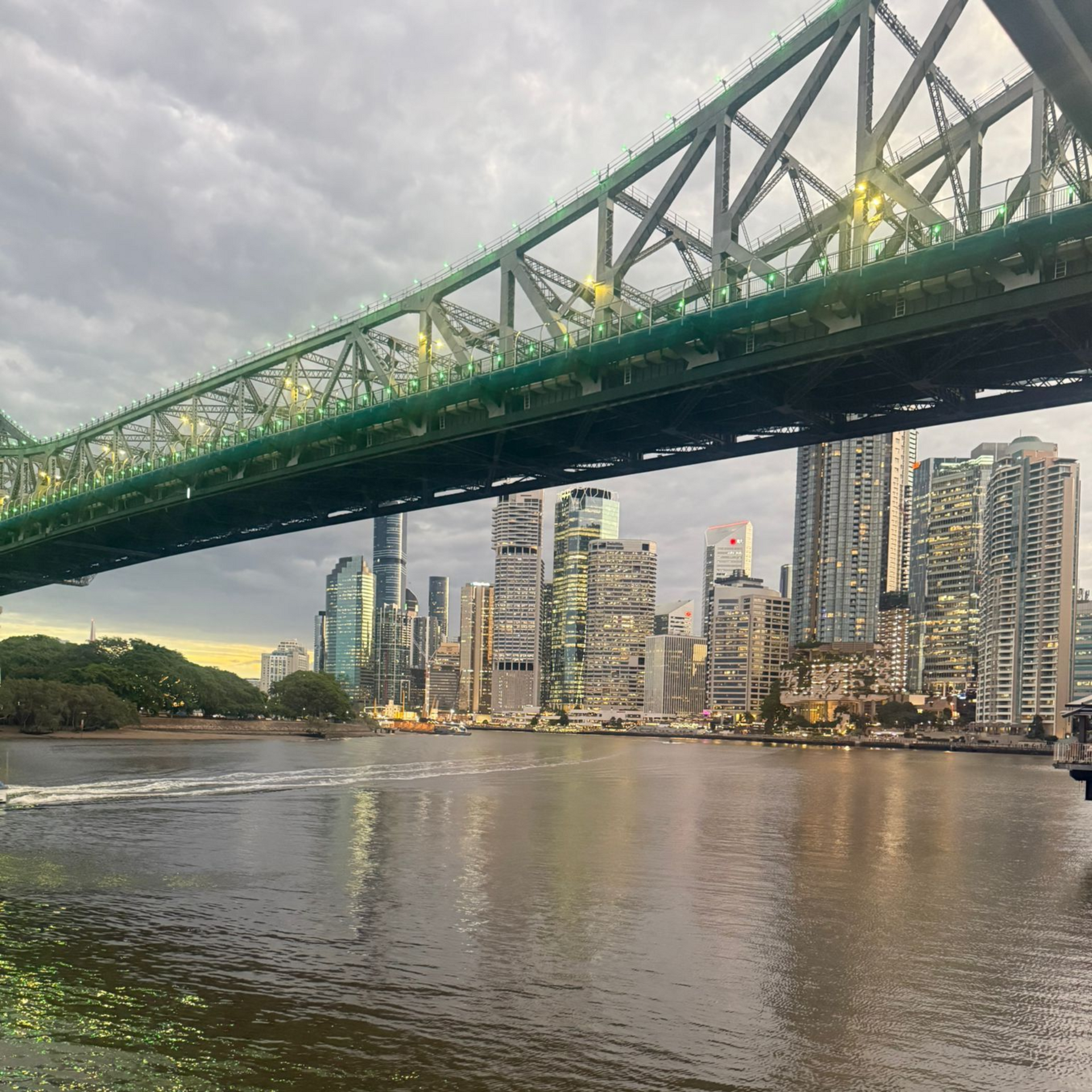 A green bridge over a river, with a city skyline in the background under a cloudy sky.