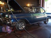 Man working on a blue vintage car with the hood open. Car is raised in a garage.