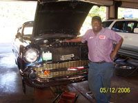 Mechanic smiles next to a classic black car with its hood open in a garage.