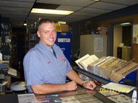 Man in blue shirt smiles behind a counter in a store, holding his arms on the counter.