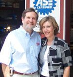 Man and woman smiling by an ASE Certified sign. Man with beard, blue shirt; woman, gray shirt, white undershirt.