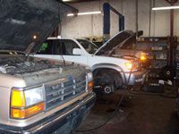 Two vehicles in a garage with open hoods. One on a lift, being worked on.