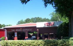 The Brake Experts auto repair shop. Red building with a sign, cars in bays, trees, and blue sky.