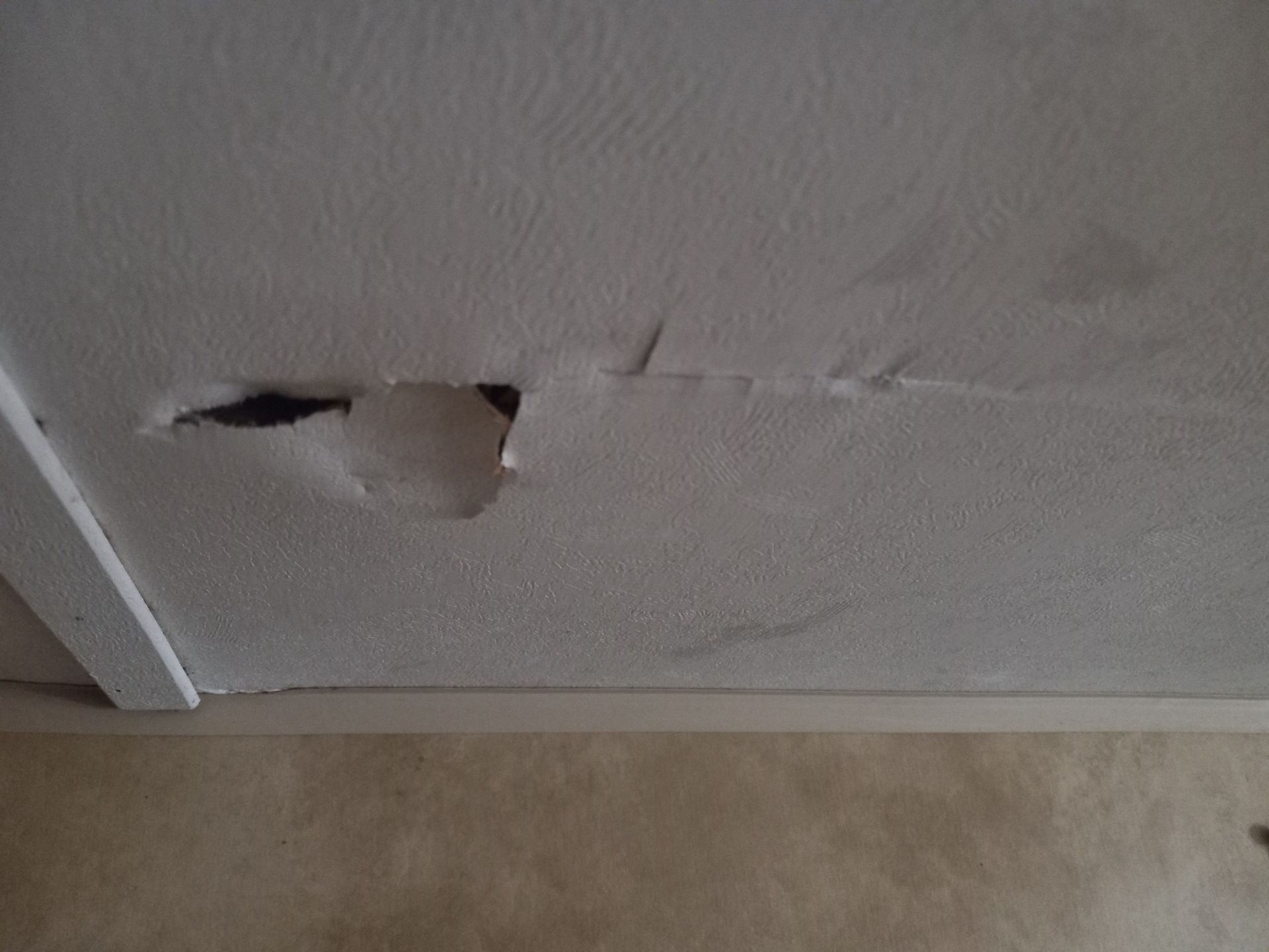 A damaged white ceiling with a large, peeling hole in the plaster near a corner trim piece.