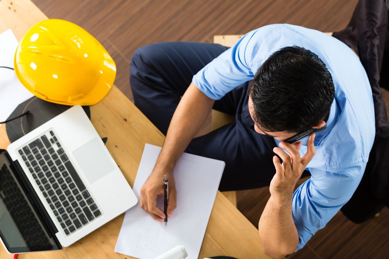 Un homme en chemise bleue, au téléphone, écrivant sur du papier à un bureau avec un ordinateur.