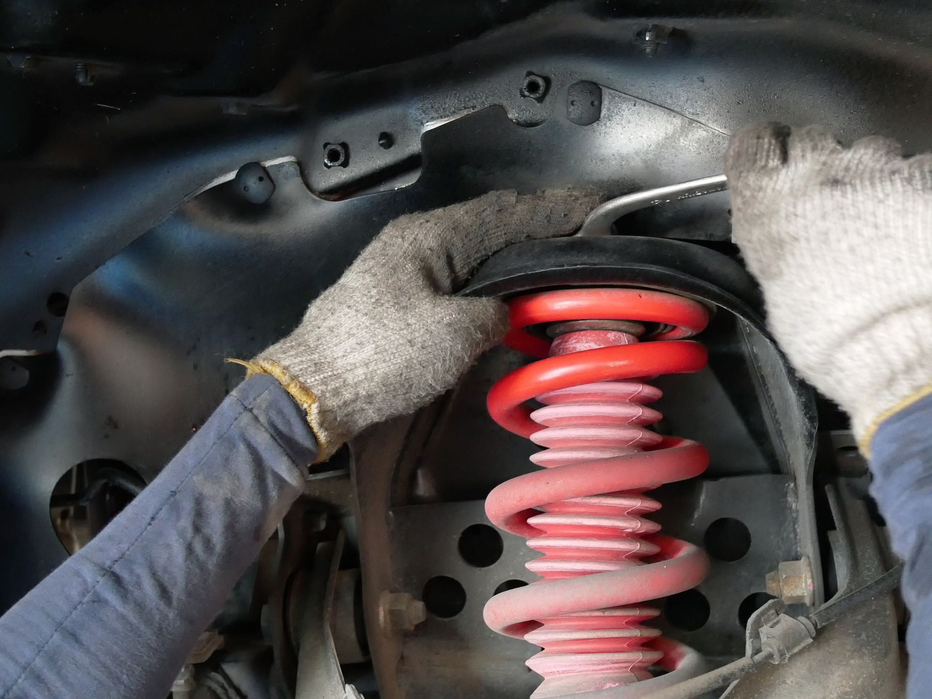 Hands in gloves working on a red car coil spring and shock absorber under the hood.