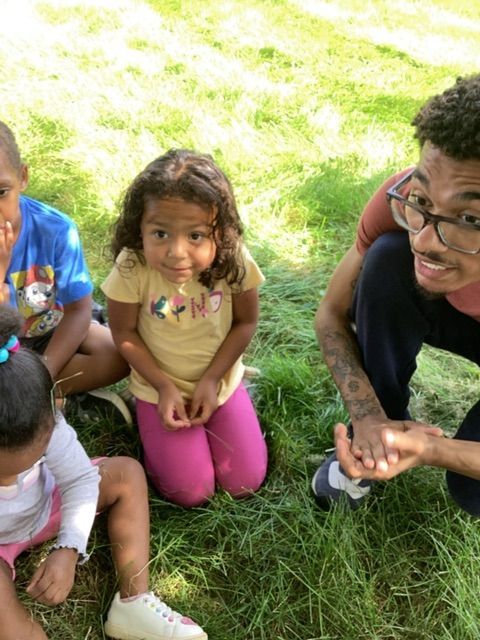 A group of children are sitting on the grass with a man kneeling down.