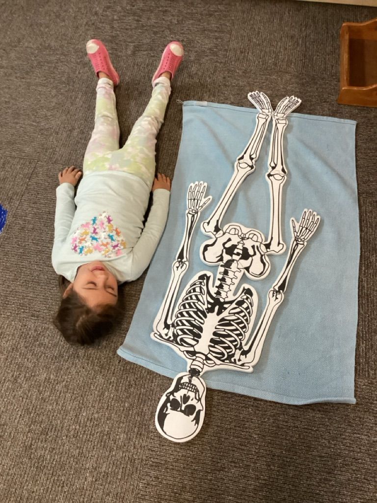 A little girl is laying on her back next to a skeleton on a towel.