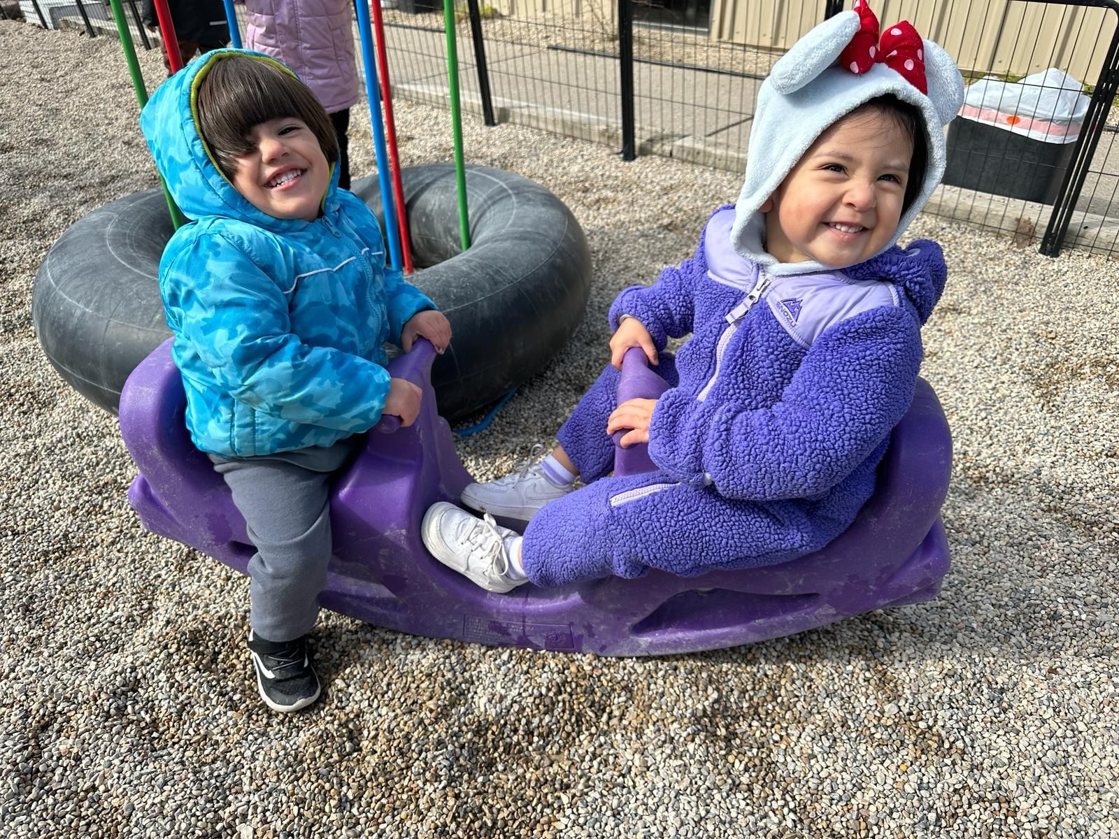 Two little girls are sitting on a purple rocking horse at a playground.