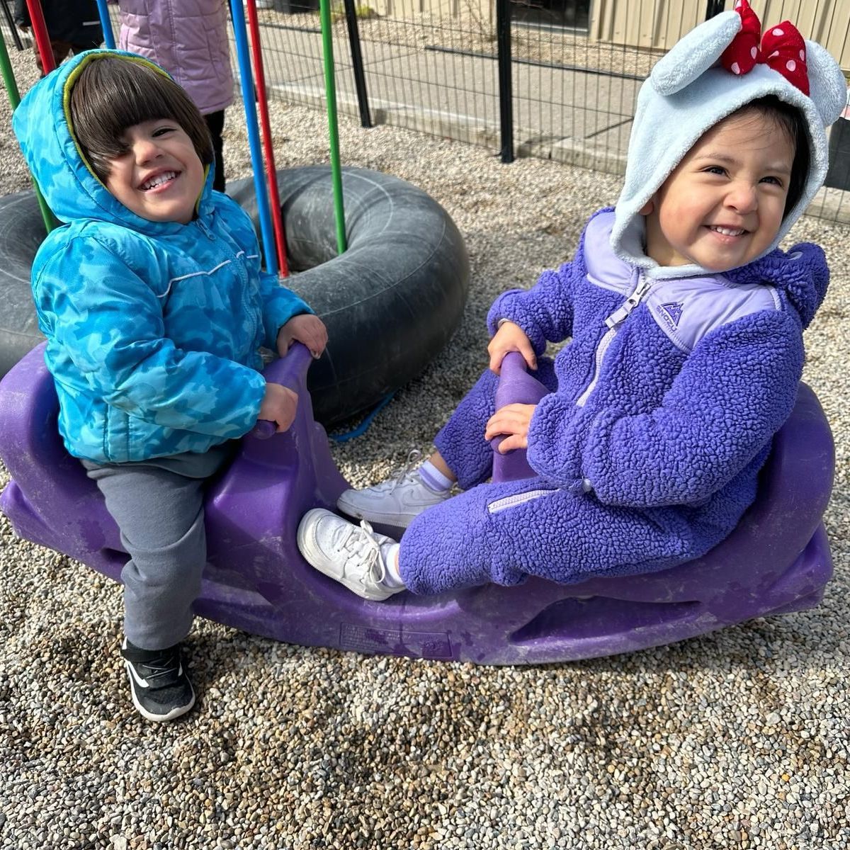 Two little girls are sitting on a purple rocking horse at a playground.