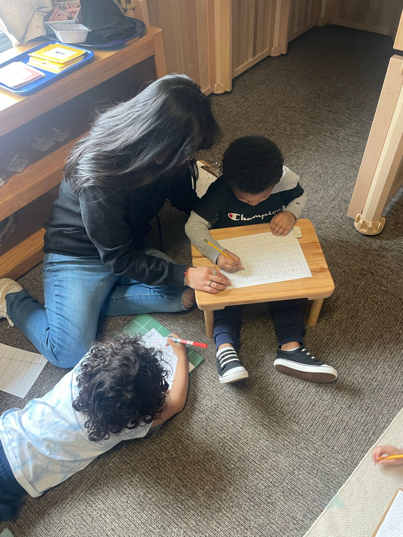 A woman is kneeling on the floor helping two children with their homework.