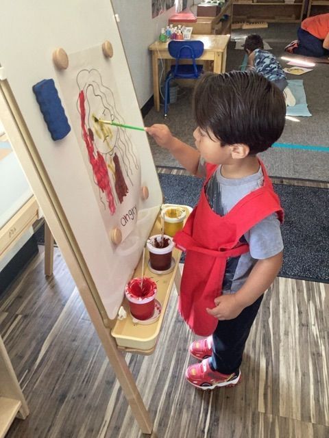 A young boy in a red apron is painting on an easel