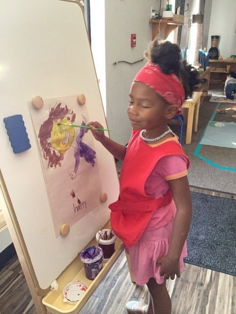 A little girl in a red apron is painting on a white board