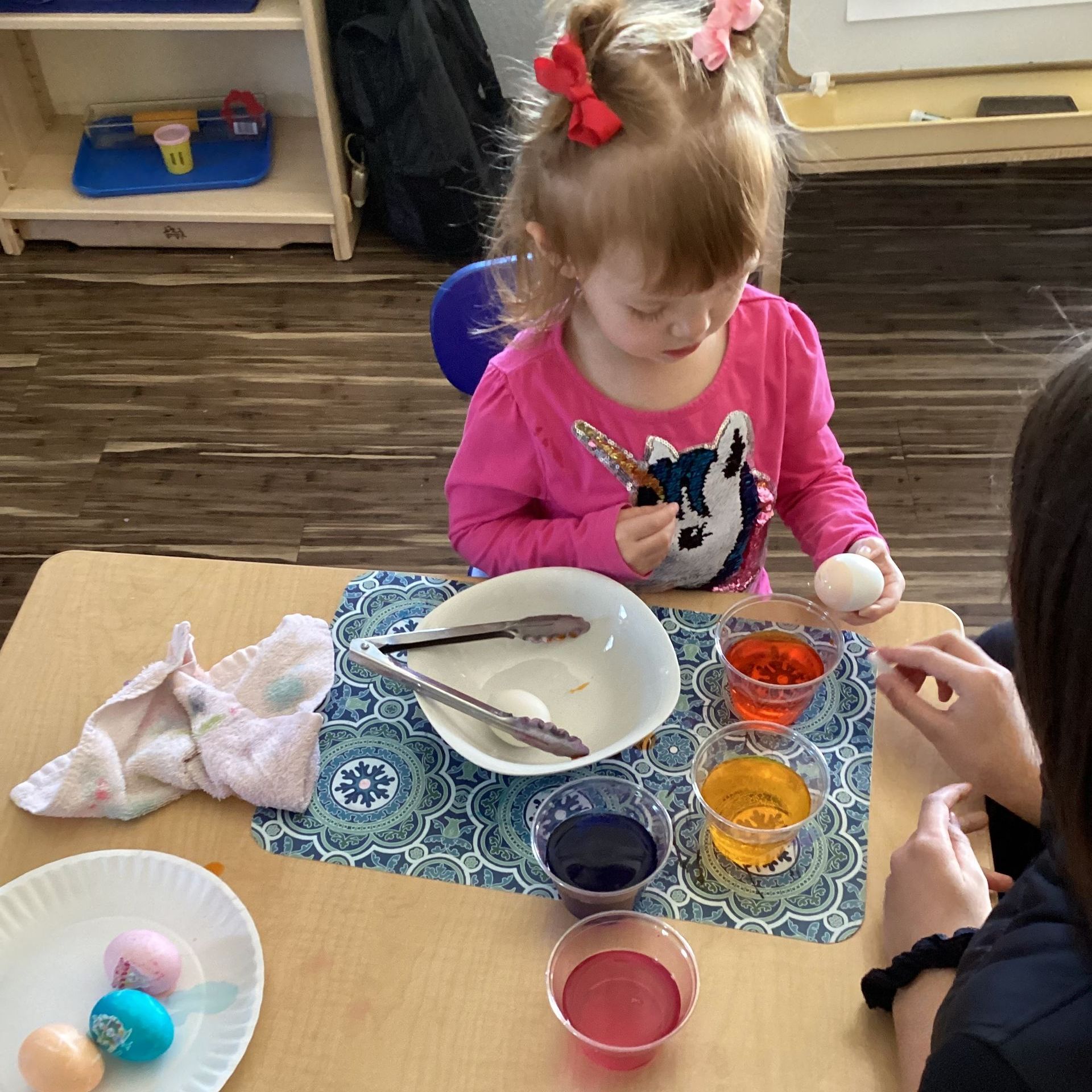 A little girl in a pink shirt is sitting at a table holding an egg