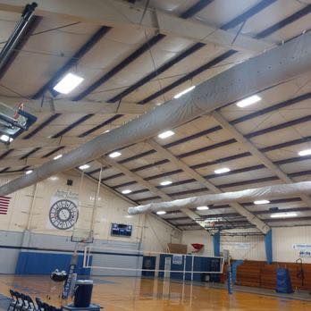 Gym interior with a volleyball net, blue walls, and a high, sloped ceiling with lights and air ducts.