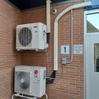 Two white air conditioning units mounted on a brick wall near a door labeled 