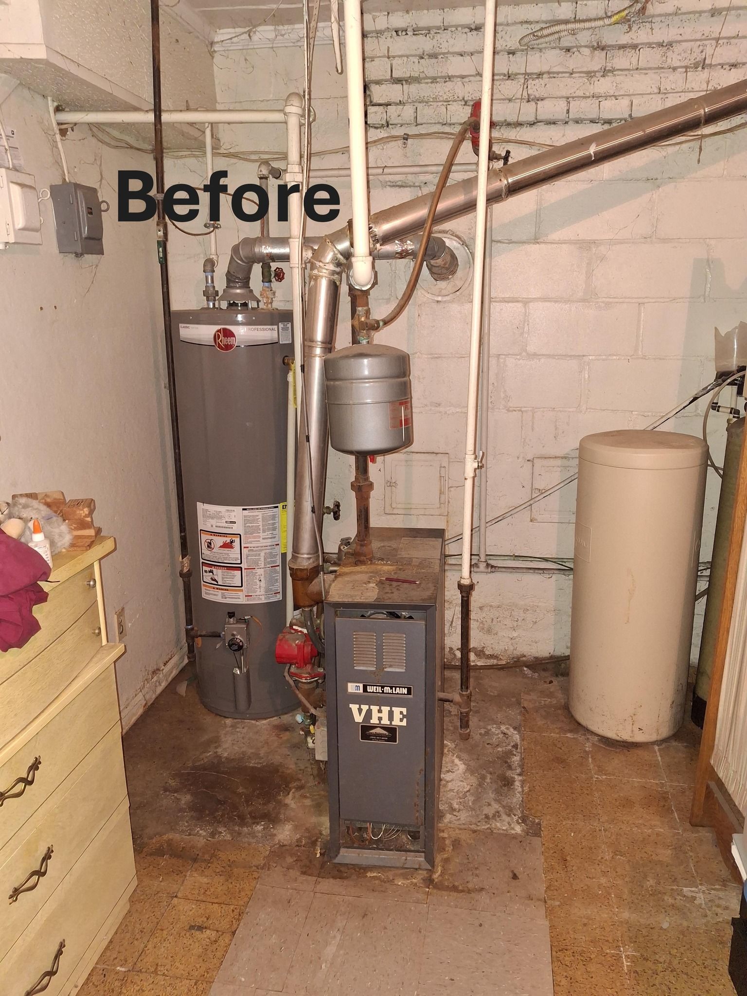 Basement with water heater, furnace, and tank. Worn surfaces, pipes, and exposed brick walls. 