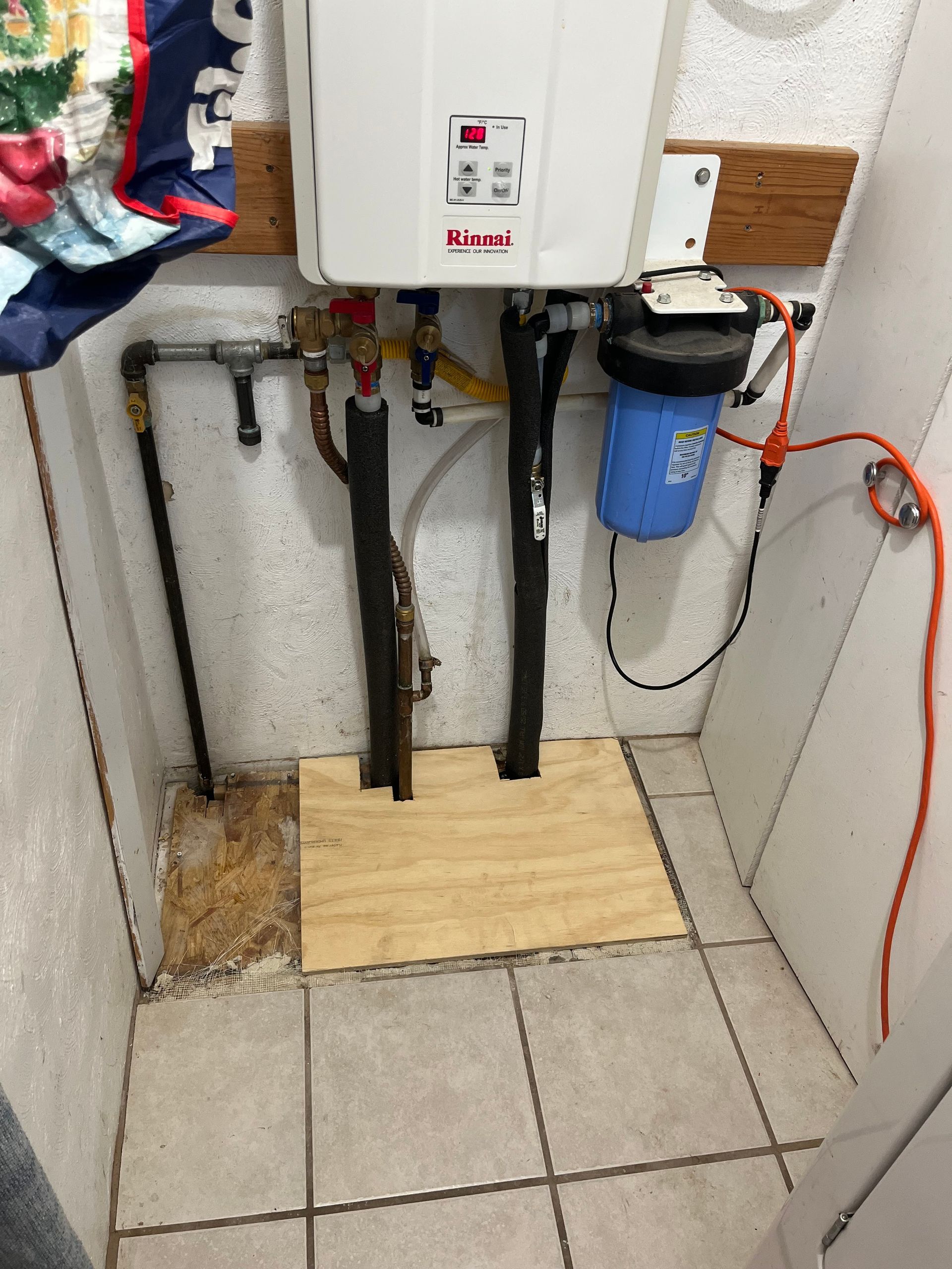 White tankless water heater, with filter and plumbing, mounted on wall in tiled utility room.