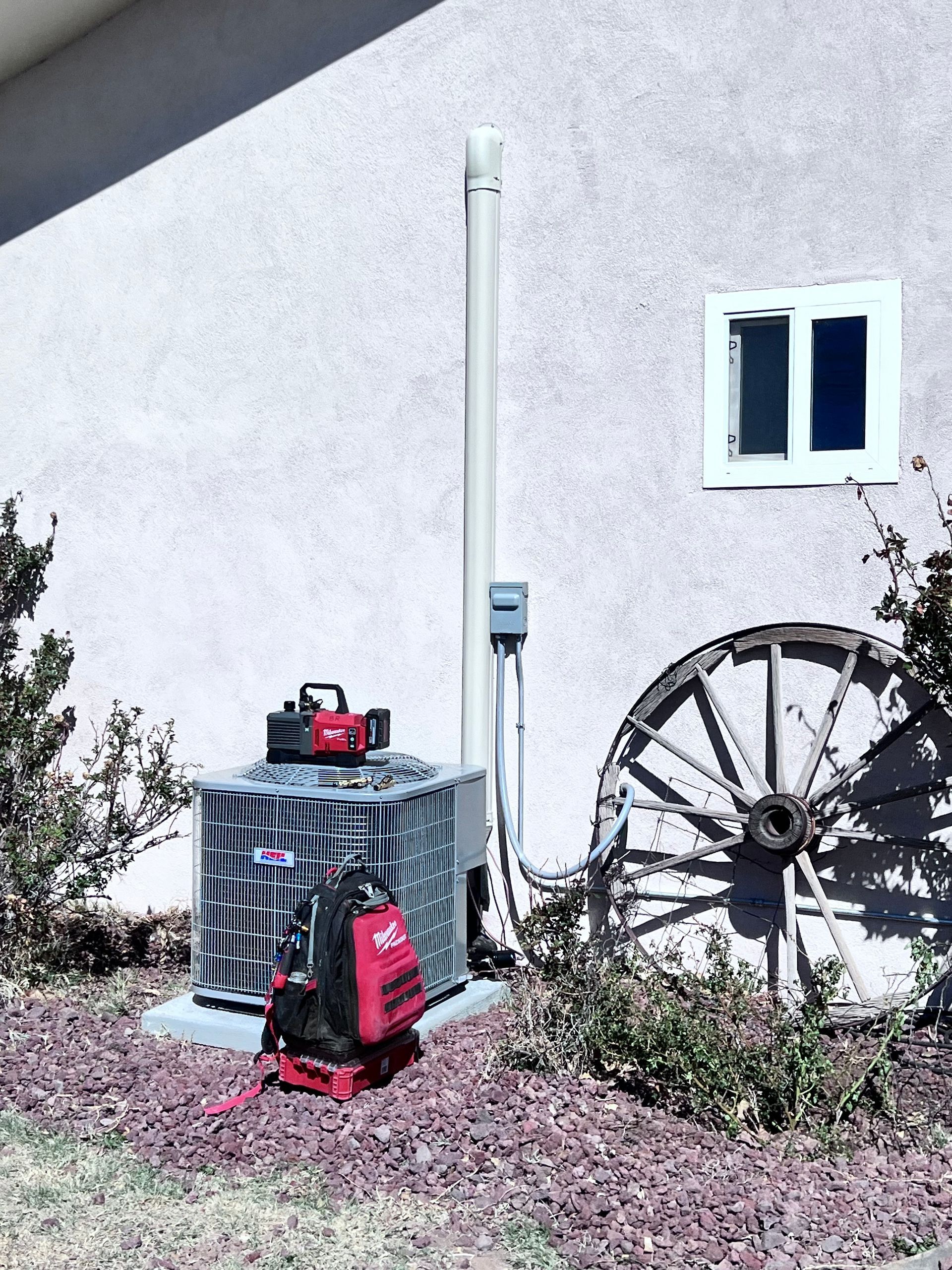 HVAC unit with tools, electrical box, and wagon wheel by stucco wall, next to greenery.
