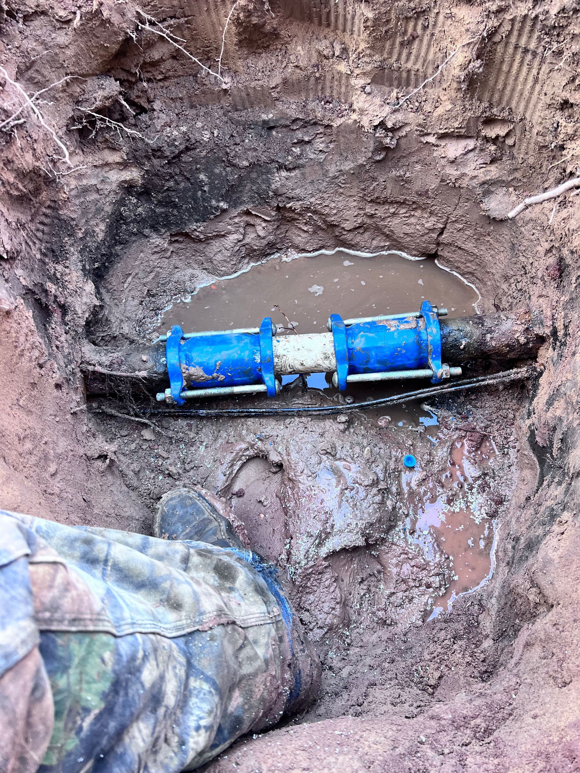 Blue-jointed pipe in a muddy hole. A leg in camo boot is present. Water pools around the pipe.