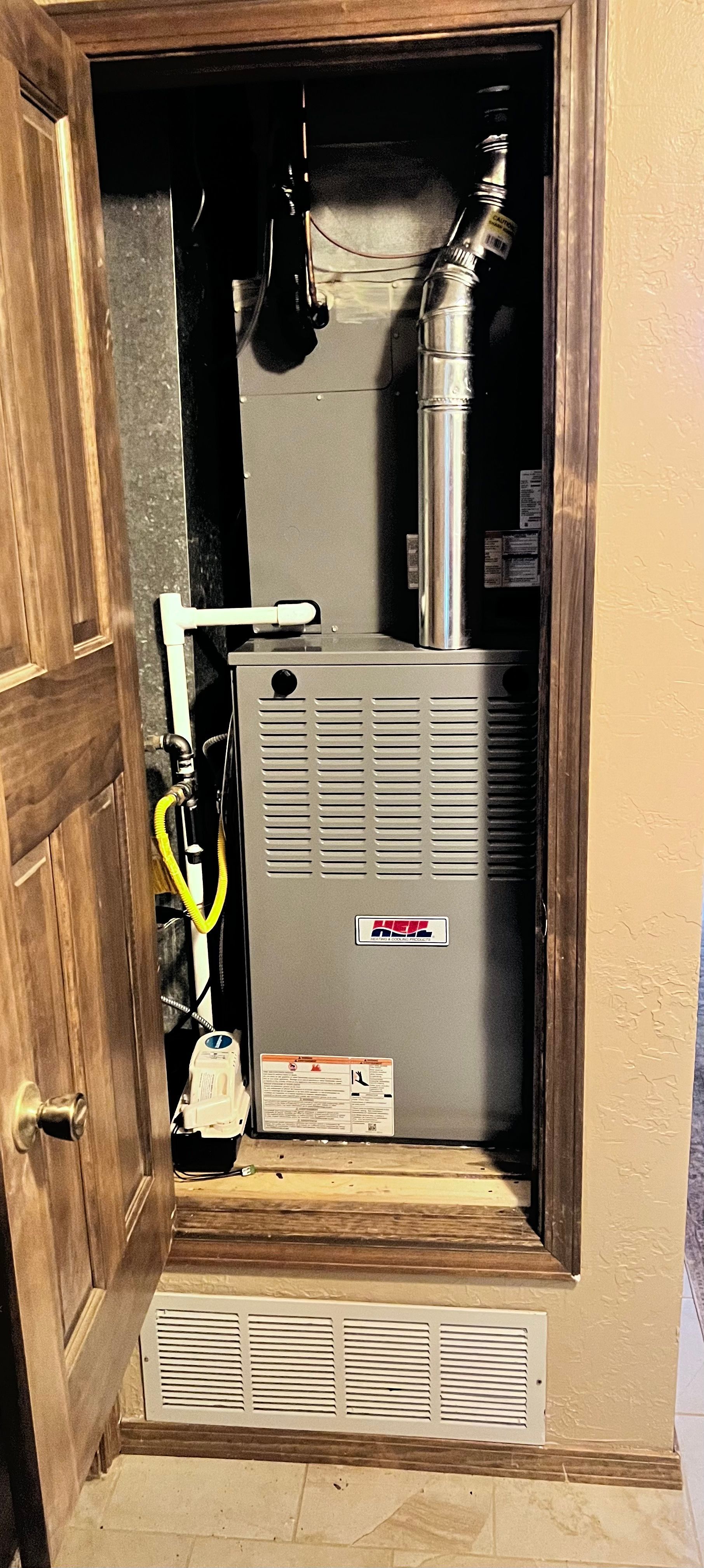 Furnace inside a wooden-framed closet; door open to the left, vent below furnace, pipes above.