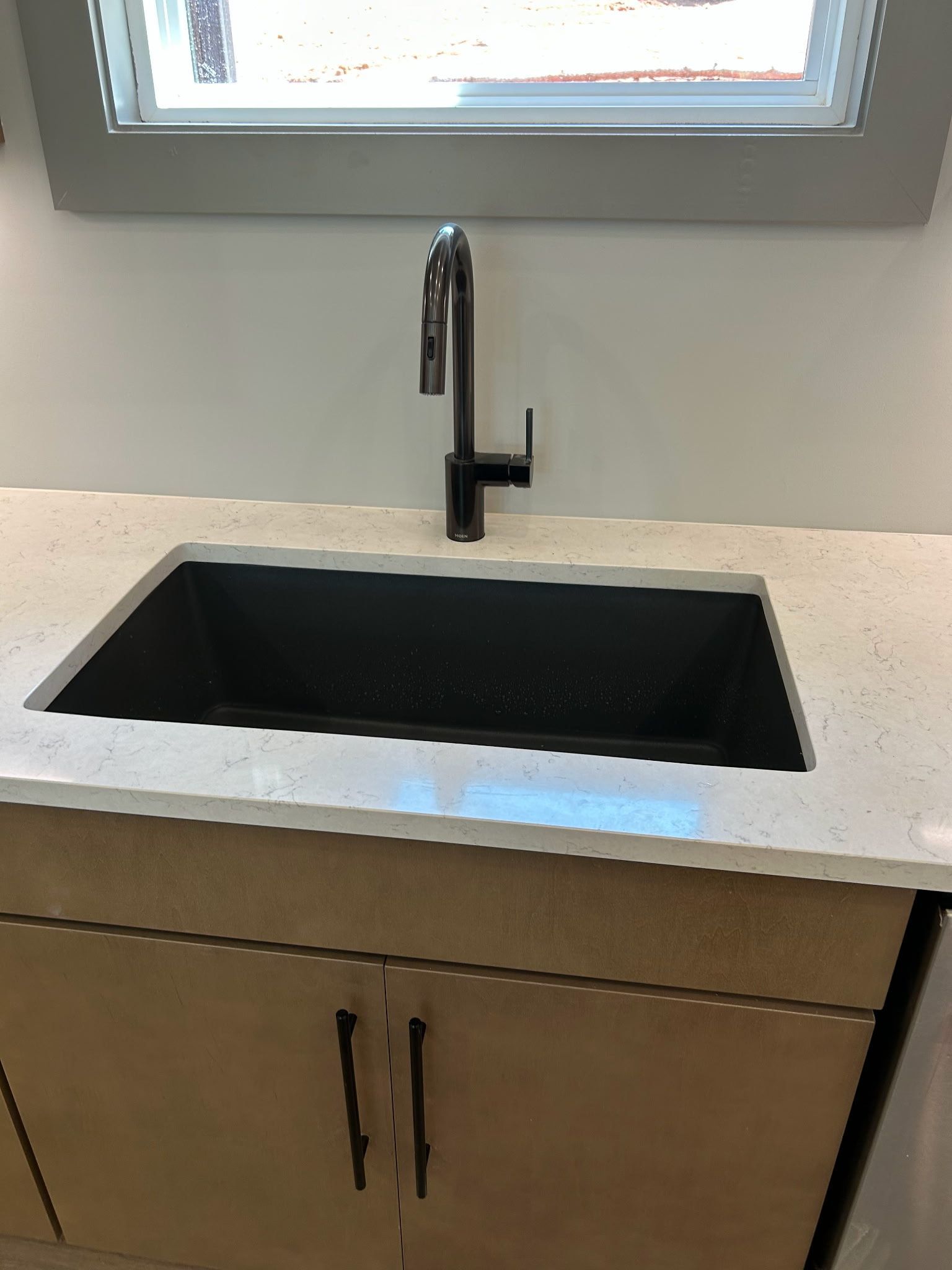 Black sink with dark faucet on a light countertop and light wood cabinets under a window.