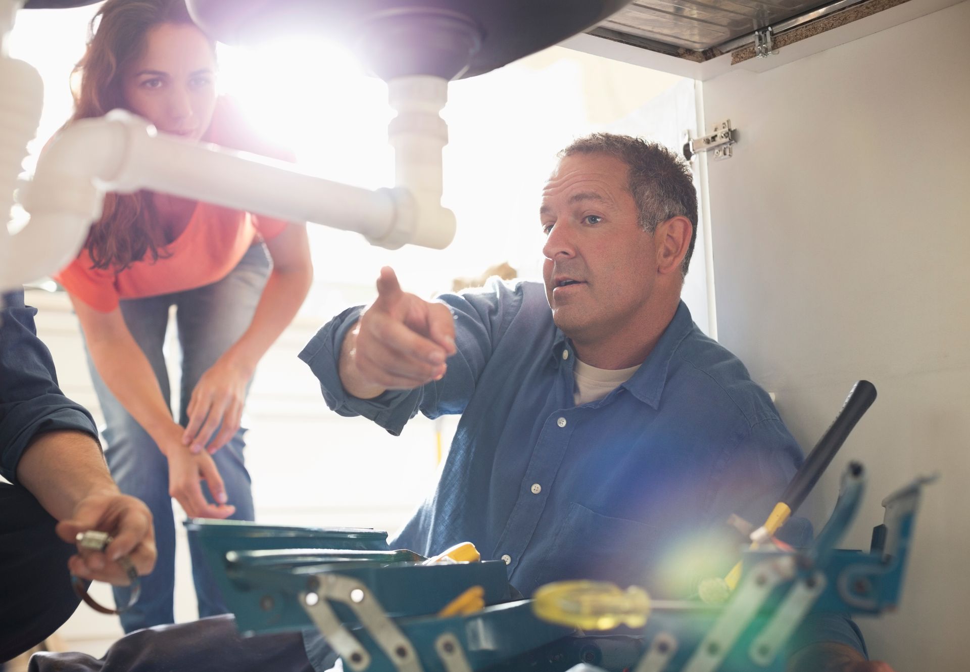 A plumber under a sink with a homeowner, repairing a kitchen sink while explaining.