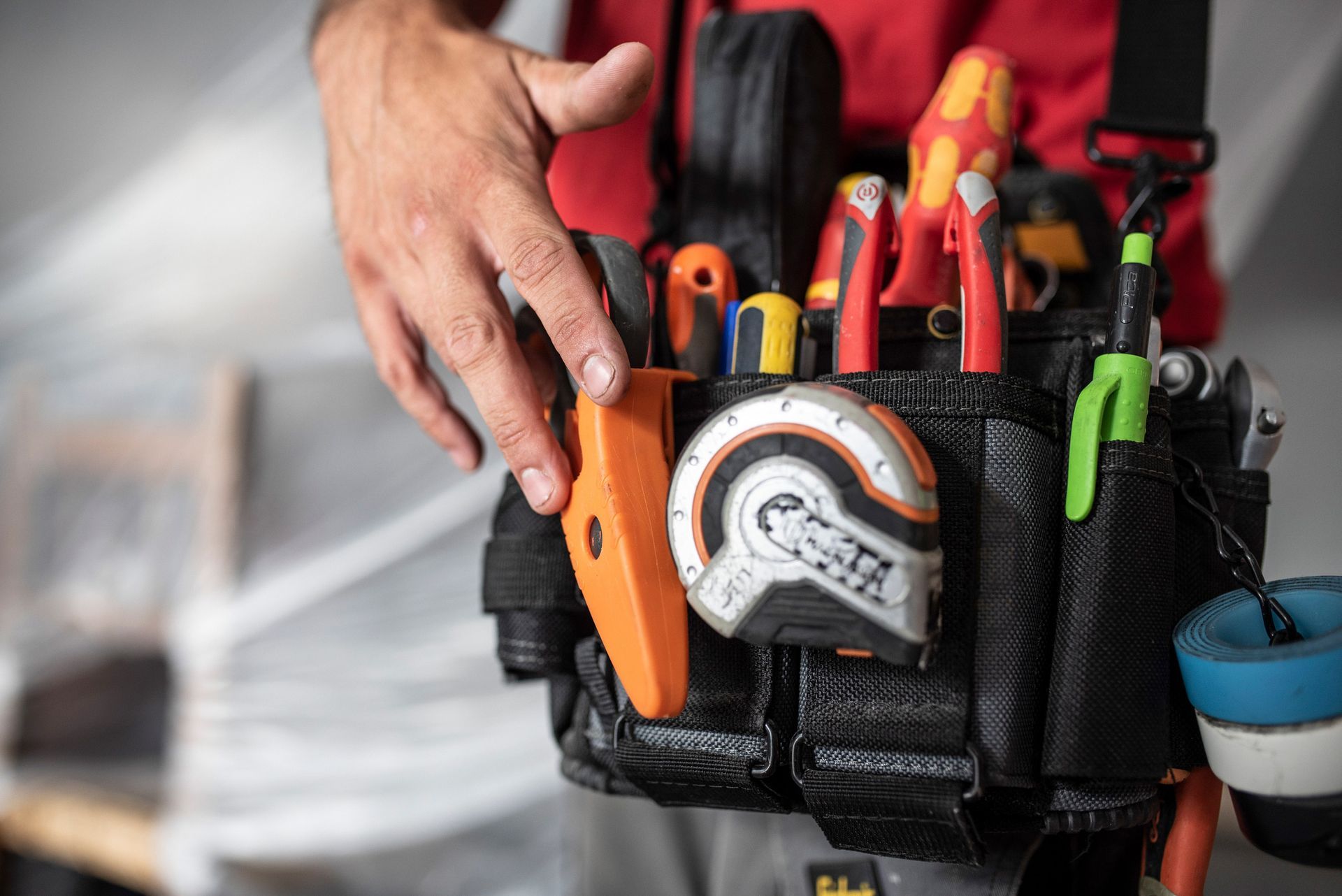 Hand reaching for an orange tool in a black tool belt. Electrical tools and a tape measure are visible.