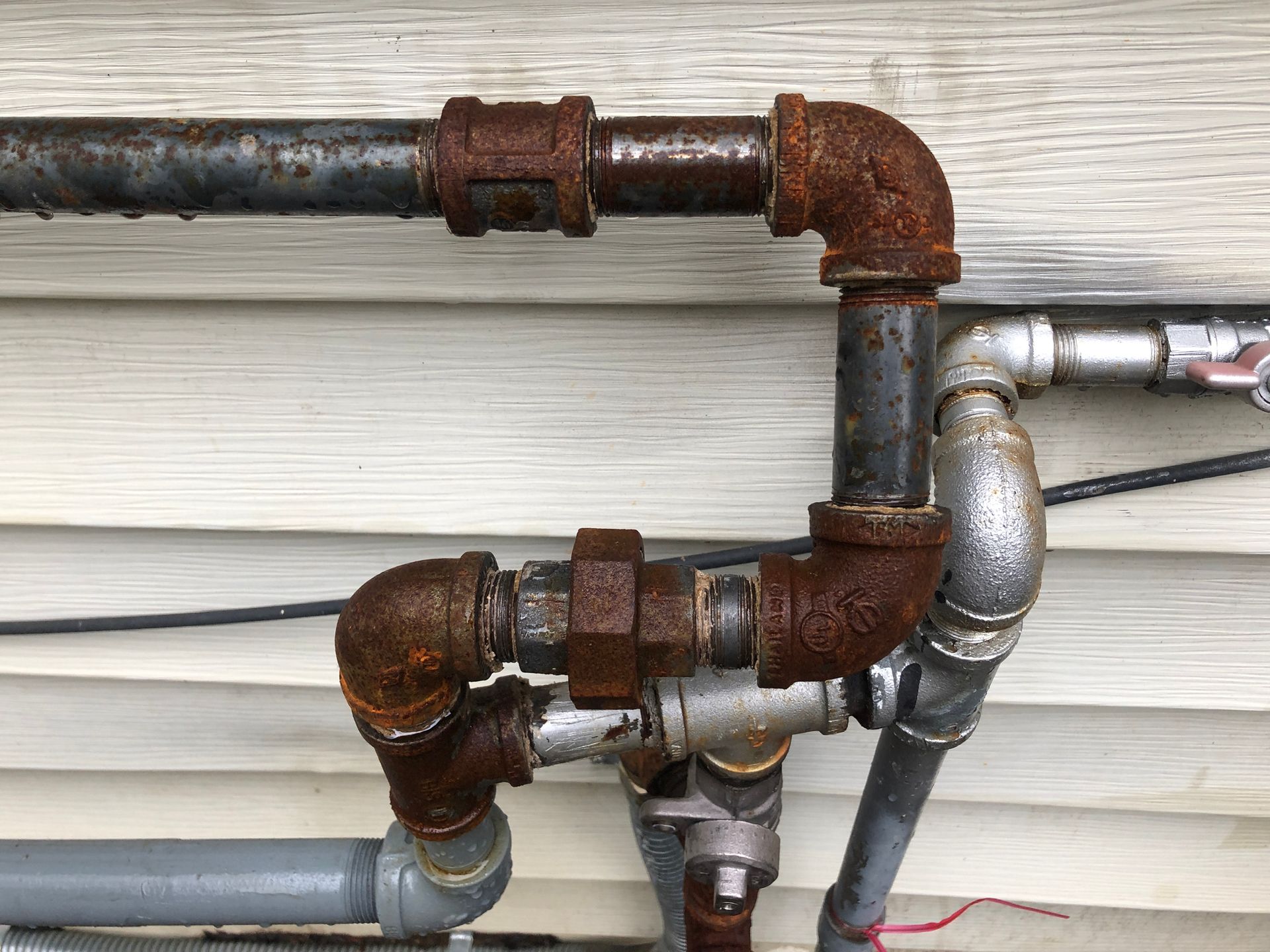 Rusty metal pipes with various elbows and fittings, connected to a wall.