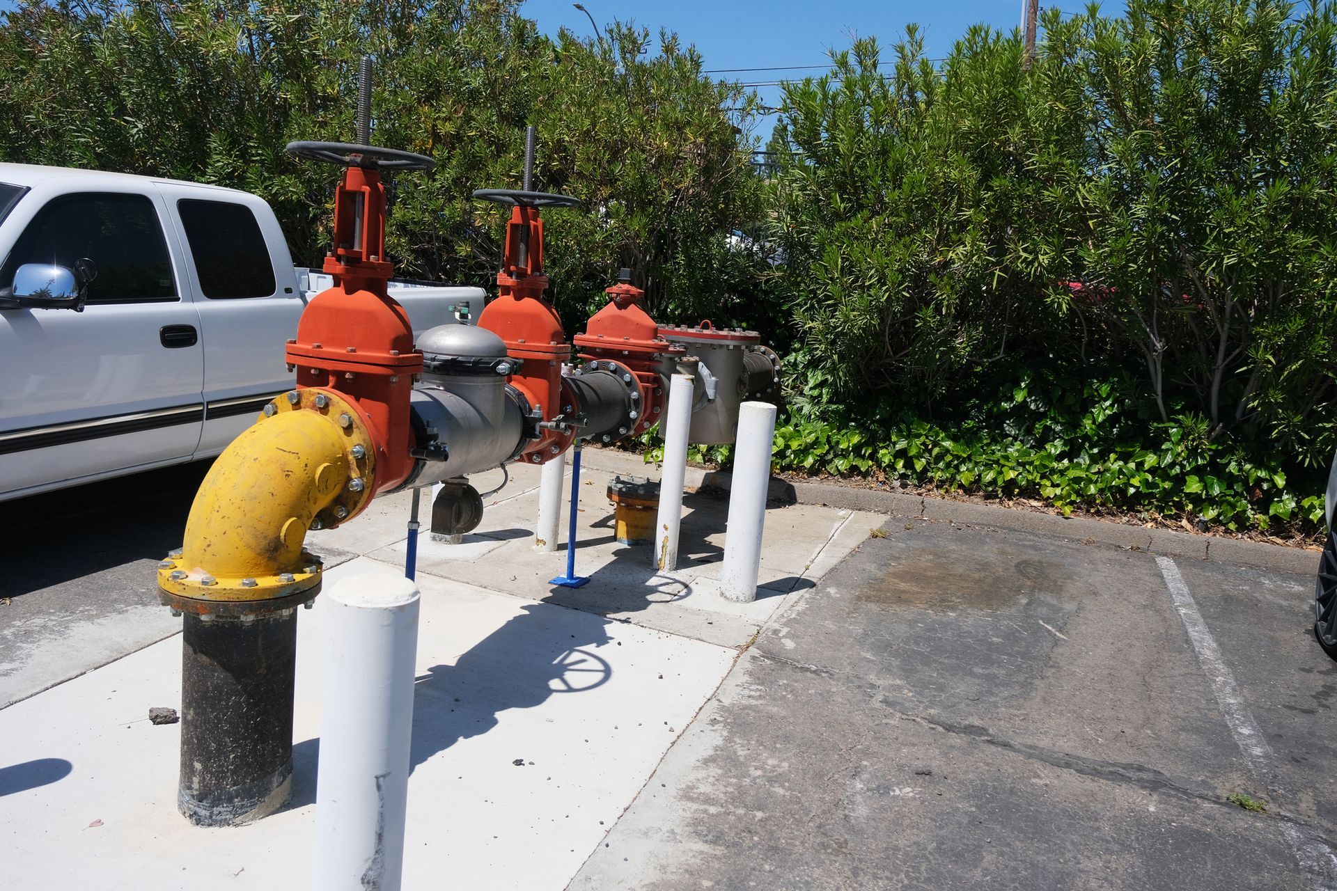 Water valves with red and yellow pipes on a concrete slab, next to a white truck and green bushes.